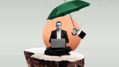 A businessman sits cross-legged on a floating rock, working on a laptop under a green umbrella.