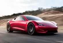 A sleek red Tesla Roadster driving dynamically along a paved coastal road with blurred background hills.