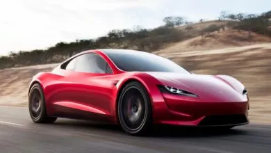 A sleek red Tesla Roadster driving dynamically along a paved coastal road with blurred background hills.