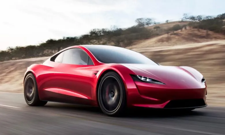 A sleek red Tesla Roadster driving dynamically along a paved coastal road with blurred background hills.