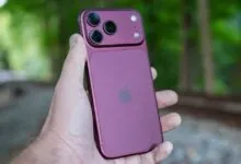 A person holds a dark purple Apple iPhone with a prominent triple-camera module outdoors.