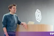 Sam Altman leaning on a podium in front of a grey wall with the OpenAI logo