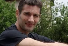 Close-up portrait of a smiling man outdoors with green foliage in the background.