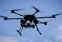 A black hexacopter drone in flight against a bright blue sky with soft white clouds.