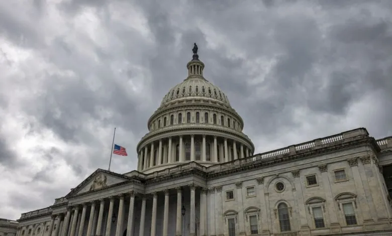 The US Capitol building stands under a dramatic, cloudy grey sky with an American flag flying at half-mast.