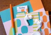 White AirPods rest on colorful patterned notebooks beside an orange pen on wood.