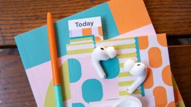 White AirPods rest on colorful patterned notebooks beside an orange pen on wood.