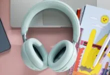 Mint green wireless headphones rest on a pink desk next to a smartphone, a laptop, and colorful notebooks.