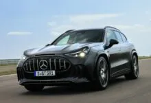 A dark grey Mercedes-AMG SUV driving fast on a paved test track under a bright, partly cloudy sky.