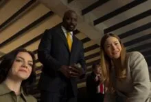 Three people smiling while looking down, featuring actor Mike Colter in the center wearing a dark suit.