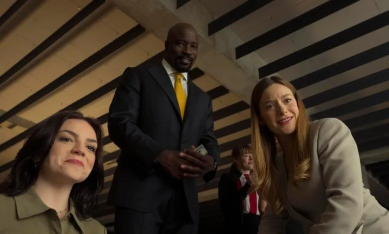 Three people smiling while looking down, featuring actor Mike Colter in the center wearing a dark suit.