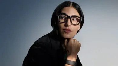 Close-up portrait of a woman wearing thick black Ray-Ban glasses and dark clothing against a gradient blue background.