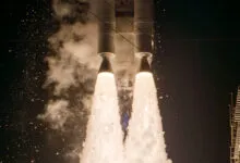 Close-up view of a rocket launching at night with massive twin plumes of bright white fire and smoke.