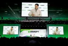 TechCrunch Disrupt conference stage with a speaker on three large screens and an audience in front