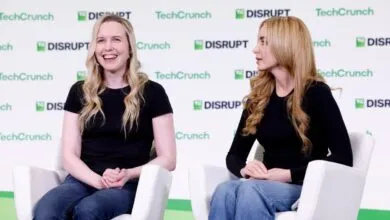 Two women seated on white armchairs at TechCrunch Disrupt stage, smiling and talking.