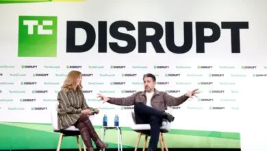 Two speakers sit on white armchairs on stage at TechCrunch Disrupt, with a large branded backdrop.
