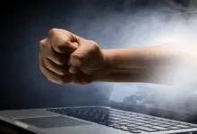 A clenched fist punches out from a laptop screen surrounded by smoke and dramatic lighting.