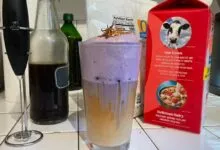 A tall glass of layered purple and white frothy coffee sits on a tiled kitchen counter.