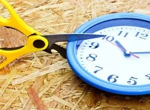 Yellow scissors cutting into the edge of a blue analog clock placed on a wooden surface.