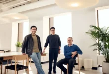 Three male founders standing and sitting together in a modern, brightly lit office space.