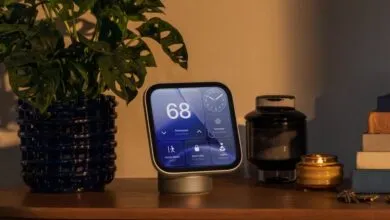 A smart home display sits on a wooden table beside a potted plant, dark jar, and lit candle.