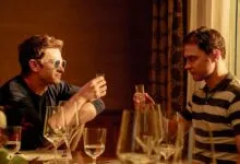 Two men seated at a restaurant table raise small shot glasses in a toast, bathed in warm indoor lighting.