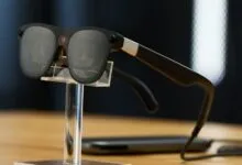 Sleek black smart glasses displayed upright on a clear acrylic stand against a dark background.