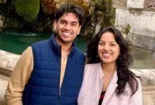 Two smiling young adults standing together outdoors in front of an old stone fountain.