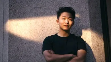 A young Asian man wearing a black t-shirt stands with arms crossed against a textured concrete wall.