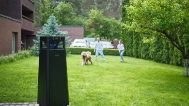 A black mosquito trap stands on a wooden deck in a lush green backyard with people and a dog in the background.