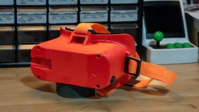 Bright orange 3D printed virtual reality headset rests on a wooden workbench in a workshop.