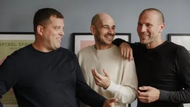 Three smiling men standing together indoors against a grey wall with framed artwork.