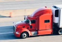 A vibrant red semi-truck pulling a white cargo trailer drives along a multi-lane highway.