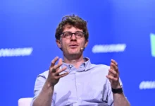 Man with glasses and striped shirt speaking on stage with blue background