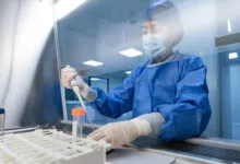 Scientist in blue protective suit uses a pipette inside a laboratory biosafety cabinet.