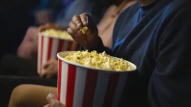 Close-up of a person reaching into a red and white striped bucket of popcorn inside a dark theater.