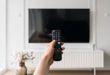 A person holds a remote control pointing toward a large wall-mounted smart television.