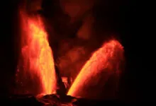 Two towering bright orange and red lava fountains erupt into the dark night sky from a volcanic crater.