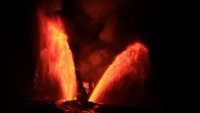 Two towering bright orange and red lava fountains erupt into the dark night sky from a volcanic crater.
