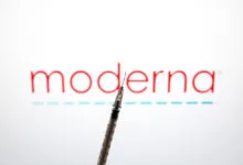 A medical syringe points upward in front of the Moderna logo on a bright screen.