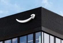 Amazon logo mounted on the dark facade of a modern corporate building against a blue sky.