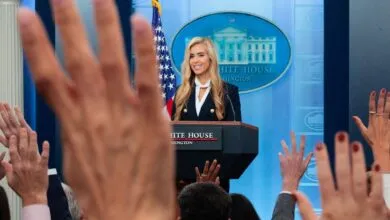 Karoline Leavitt speaks at the White House podium with raised hands visible in the foreground.