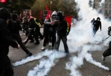 Protesters wearing face coverings advance through thick plumes of white smoke on a city street.
