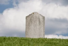 A blank weathered gray tombstone stands on a green grassy hill under a cloudy sky.
