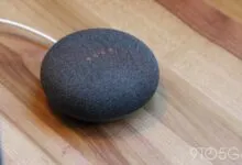 Charcoal Google Nest Mini smart speaker sitting on a wooden table with glowing LED indicator lights.
