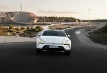 A white electric car driving on a curving asphalt road near a quarry and a body of water.