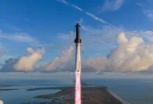 SpaceX Starship rocket launching vertically over coastal land and ocean against a blue sky with clouds.