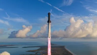 SpaceX Starship rocket launching vertically over coastal land and ocean against a blue sky with clouds.