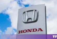 The metallic Honda logo and red lettering mounted on a white dealership sign against a cloudy blue sky.