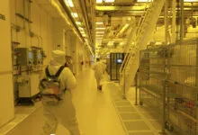 Workers in white hazmat suits walk down a sterile industrial corridor bathed in yellow light